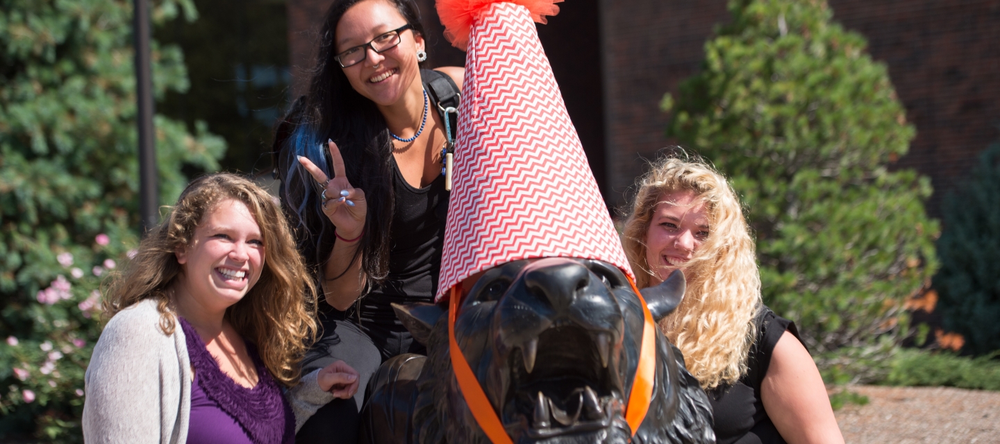 Two students standing next to tiger statue. One student is sitting on the tiger statue. 